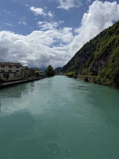 A turquoise river flows beside houses and a wooded cliff.