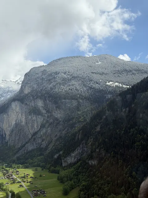 A sheer cliff and wooded mountainside tower above fields and scattered chalets.