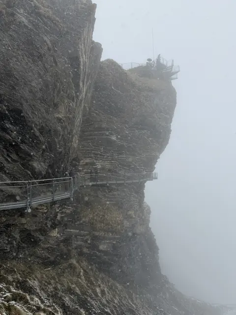 A narrow metal walkway curves around a rock face in heavy snow.