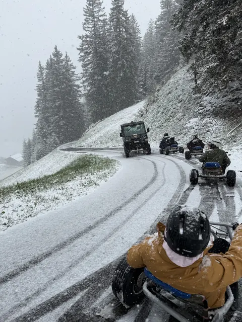Helmeted riders steer mountain carts down a snowy road lined with fir trees.
