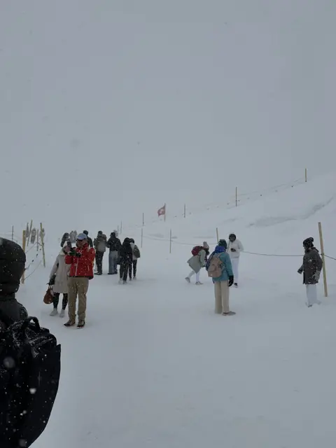 Visitors stand on a snowy mountain path with a Swiss flag in the distance.