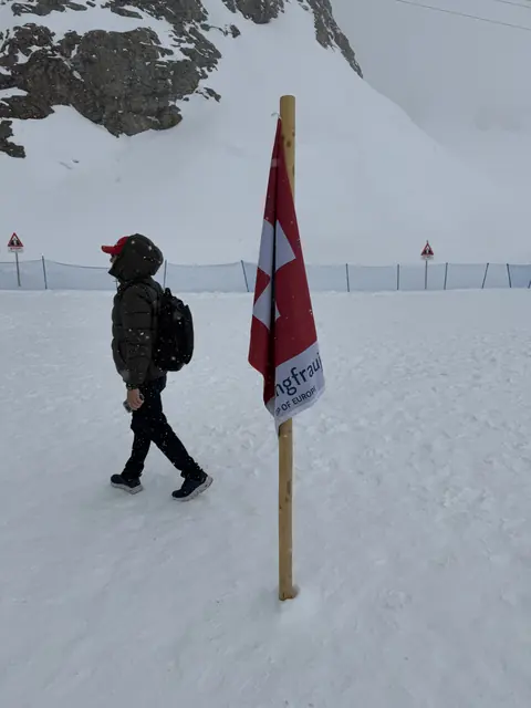 A Swiss flag on a wooden pole beside a visitor in falling snow.