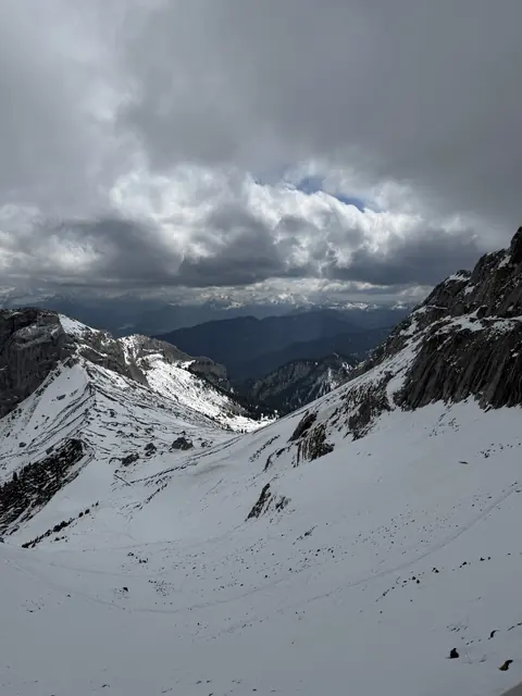 A snowy mountain basin opens toward distant ridges beneath dramatic clouds.