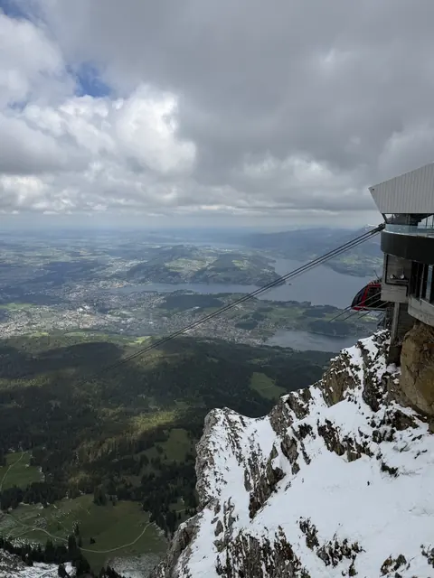 A cable car station overlooks lakes, forests, and towns from a snowy mountainside.