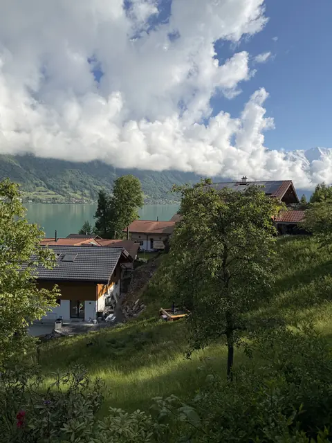 Sunlight falls on lakeside houses and green hills beneath low clouds and a blue sky.