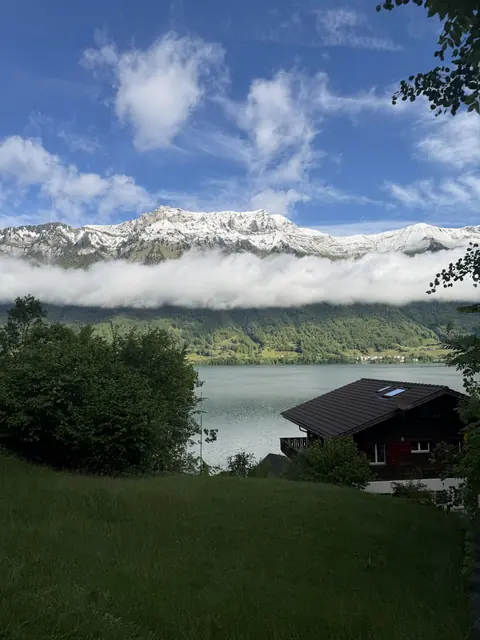 A dark wooden chalet and green lawn sit below snowy peaks and a long ribbon of cloud.