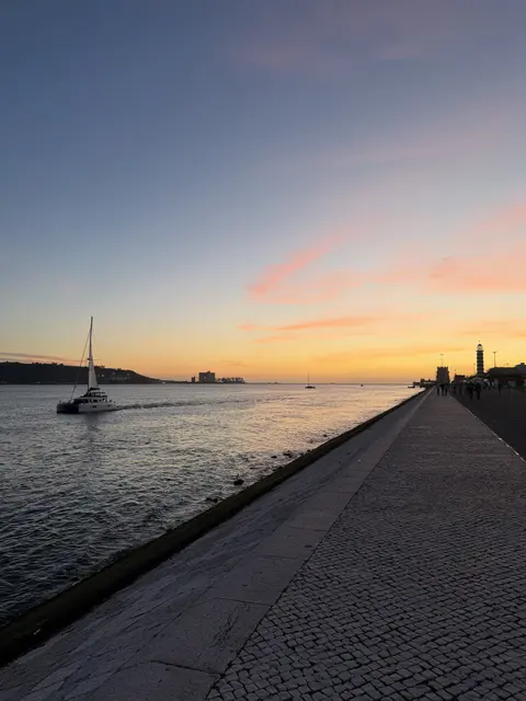 A sailboat beside a waterfront promenade at sunset.