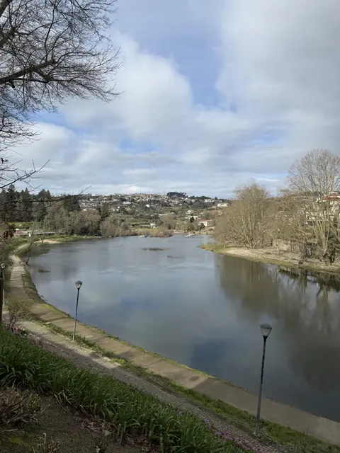 A calm river bordered by walking paths and bare trees beneath a partly cloudy sky.