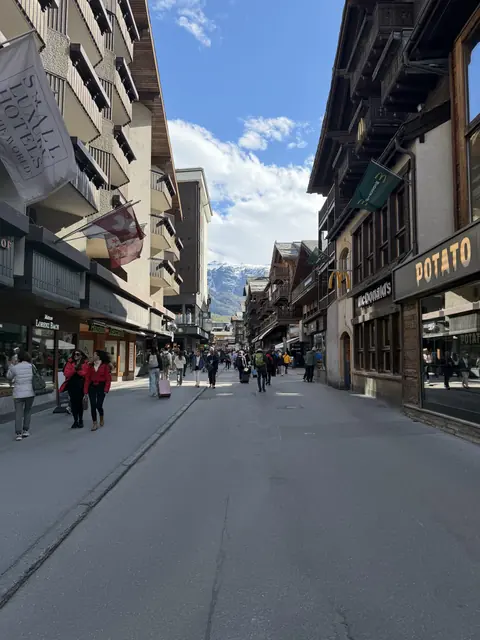 Shops and pedestrians line a village street with mountains in the distance.