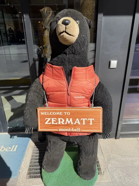 A bear wearing a red vest holds a wooden Welcome to Zermatt sign.