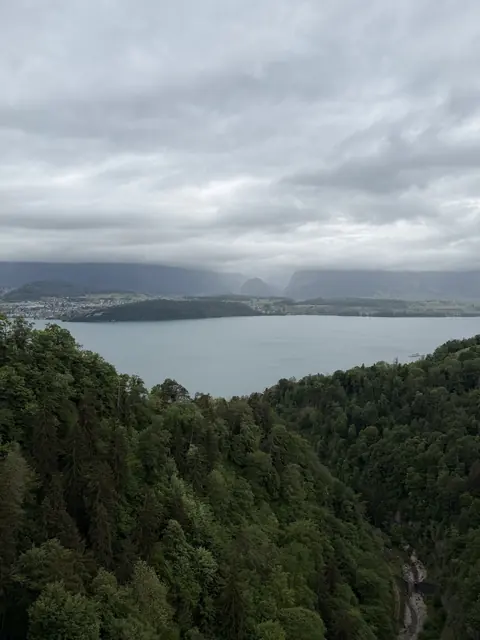 A wide lake lies beyond a forested valley beneath low clouds.